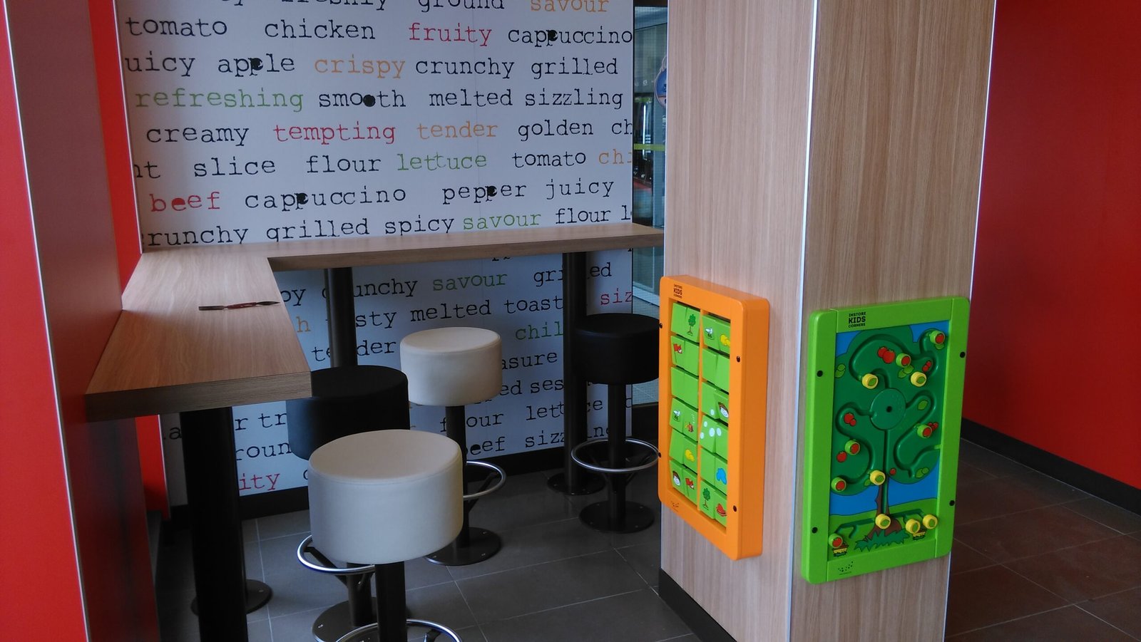 Malé posezení v restauraci rychlého občerstvení s barovými židlemi, dřevěným pultem, barevnou výzdobou stěn s nápisy o jídle a nástěnnými tabulemi s aktivitami pro děti.