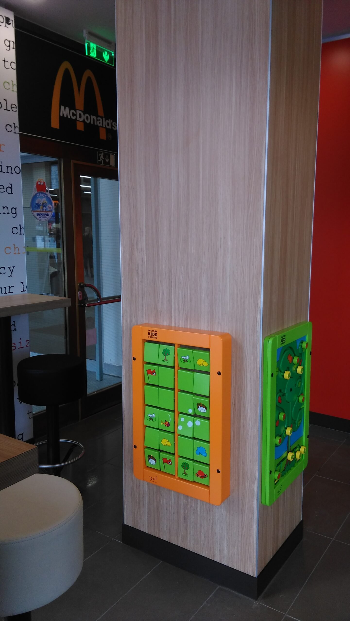 Na dřevěném sloupu v restauraci McDonalds jsou připevněny dvě tabule s dětskými aktivitami: jedna oranžová s obrázky zvířat a přírody a druhá zelená s korálky na kolejích. V pozadí je vidět vchod do McDonalds a místa k sezení.