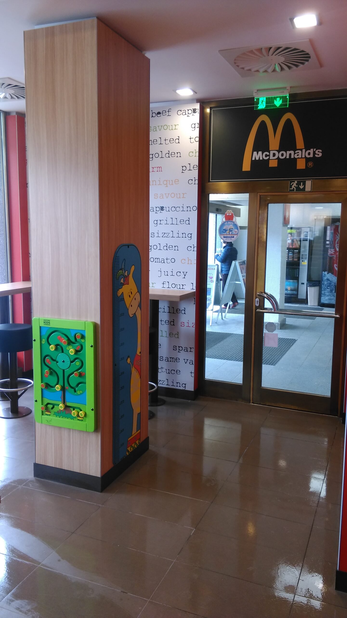 Vchod do restaurace McDonald's se skleněnými dveřmi, dřevěným sloupem s výškovou tabulkou žirafy a nástěnným korálkovým bludištěm, lesklou kachličkovou podlahou a stěnou s motivem jídelního lístku v pozadí.