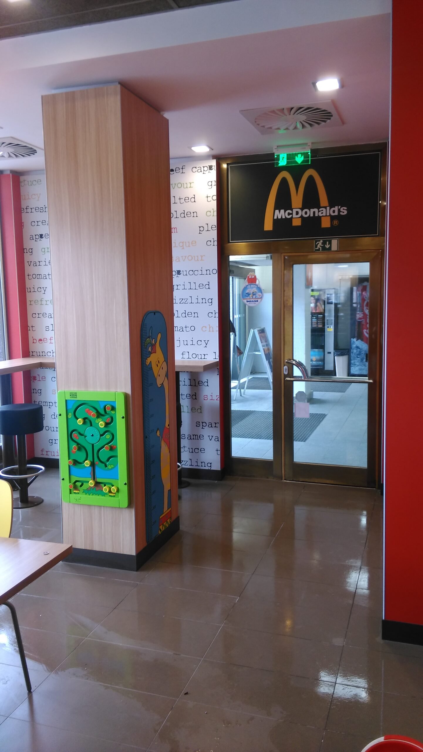 Interiér restaurace McDonald's se skleněnými dveřmi k východu, dřevěným sloupem s hravou kreslenou grafikou a stěnou s textem na pozadí. V popředí jsou vidět stoly a židle.
