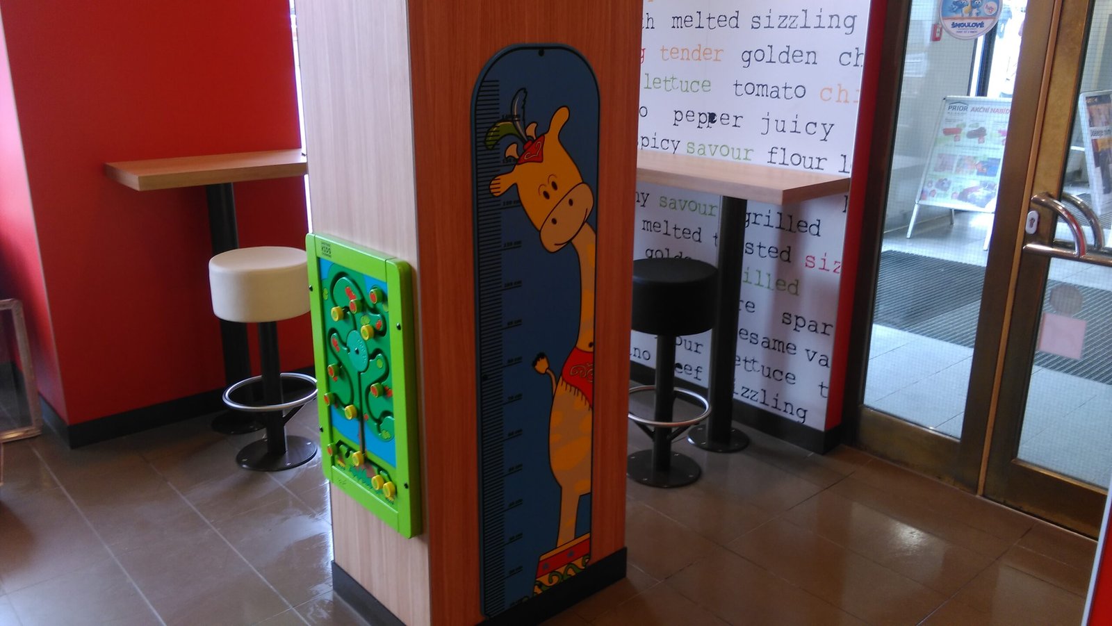 Dětská výšková tabulka s kreslenou žirafou je připevněna na dřevěném sloupu v restauraci. Vedle ní je zelený interaktivní hrací panel. V pozadí jsou barové židle a stůl.